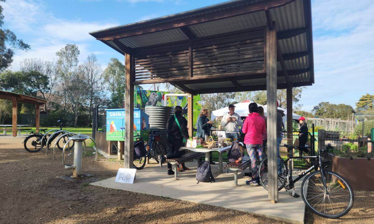 Photo of people standing around under an open-air shed. They're gathered around a picnic table, in front of a community garden.
